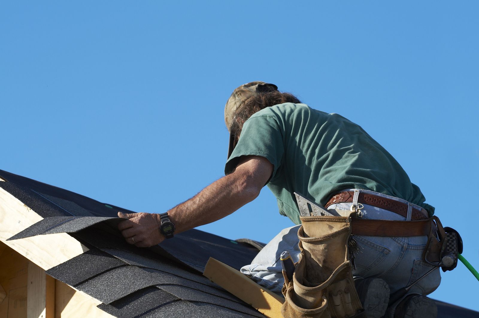 Roofer nailing shingles along the eave of a pitched roof under a clear sky